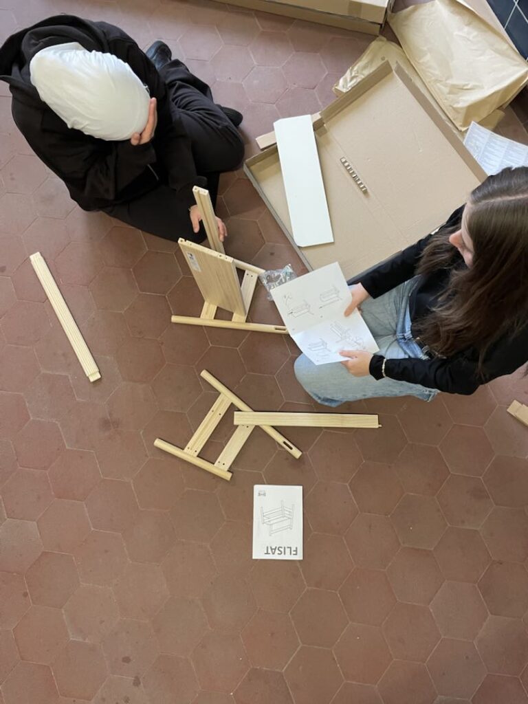 Zwei Schüler bauen am Boden mit Aufbauanleitung ein Holzmöbel zusammen