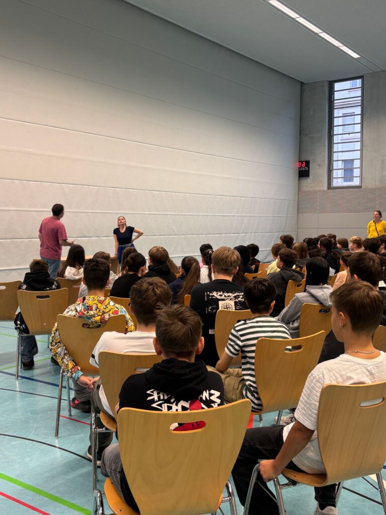 Schülerinnen und Schüler sitzen auf Stühlen in der Halle und schauen auf die beiden Schauspieler
