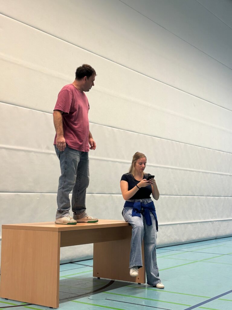 Die beiden Schauspieler auf und an einem Tisch vor der Hallenwand